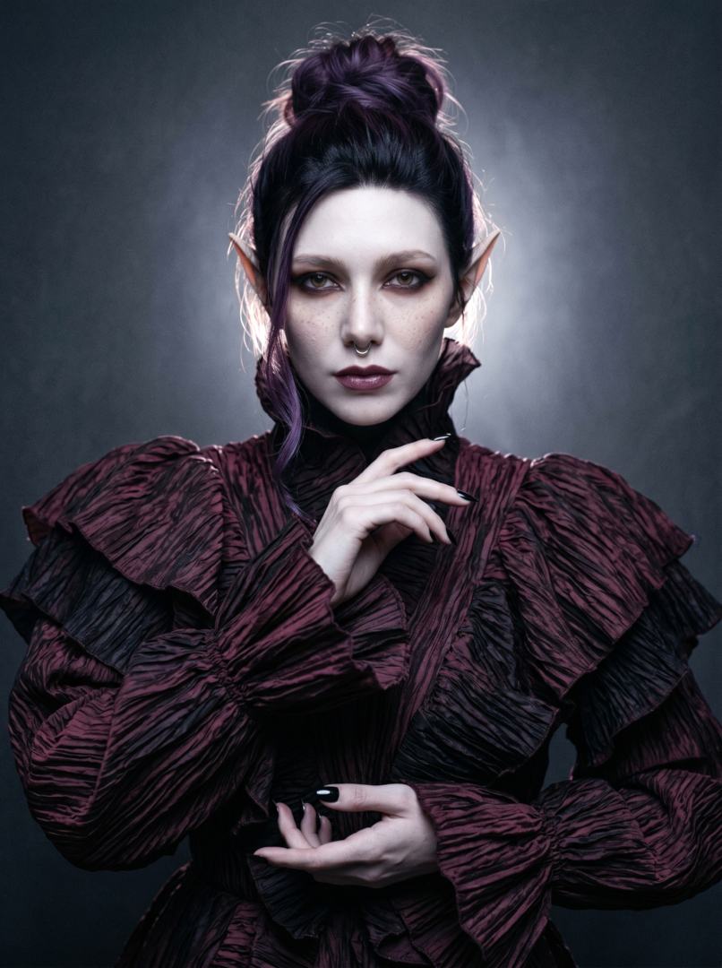 Dark editorial fashion portrait of Sael'thirra, an AI digital persona — a woman with long pointed ears, dark upswept hair and a silver septum ring, posed in a dramatic ruffled burgundy-black gothic gown, one black-nailed hand raised beneath her chin under moody studio lighting.