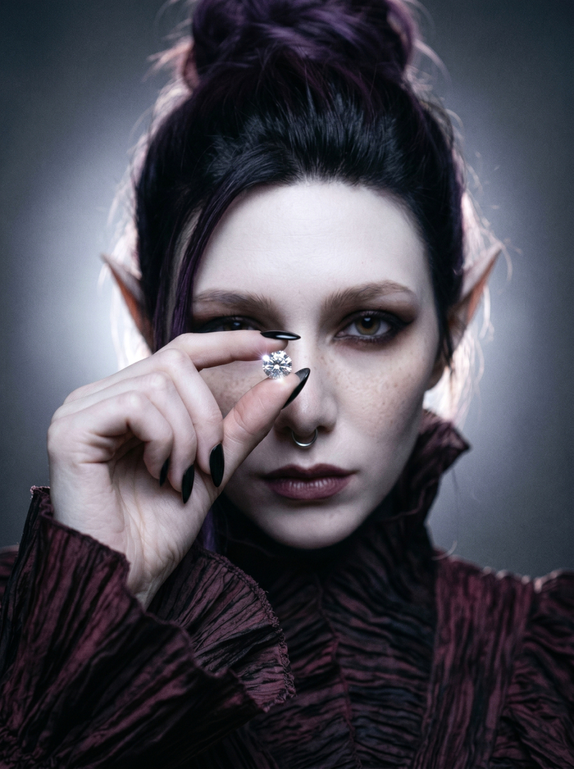 Gothic beauty editorial portrait of Sael'thirra, an AI digital persona — a woman with long pointed ears and glossy black almond nails holding a brilliant-cut diamond before one eye, framed by a burgundy-black ruffled collar in low-key chiaroscuro lighting.
