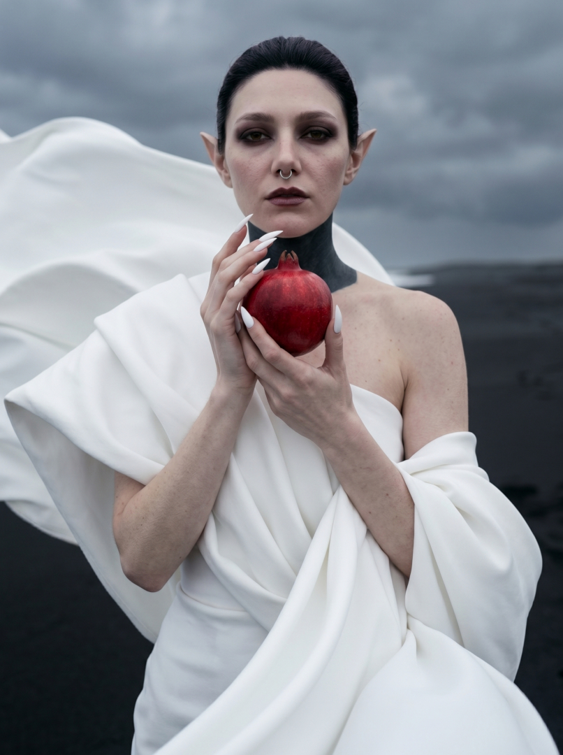 Surreal fashion editorial portrait of Sael'thirra, an AI digital persona — a woman with long pointed ears, slicked-back hair and a black-painted throat, cradling a red pomegranate in sculptural white draped fabric against a stormy grey sky, evoking a Persephone myth aesthetic.