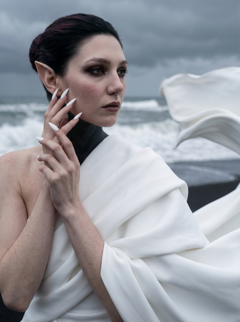 Avant-garde fashion editorial portrait of Sael'thirra, an AI digital persona — a woman with long pointed ears and long white stiletto nails in profile, draped in flowing white silk on a black-sand volcanic shore with a stormy ocean behind.
