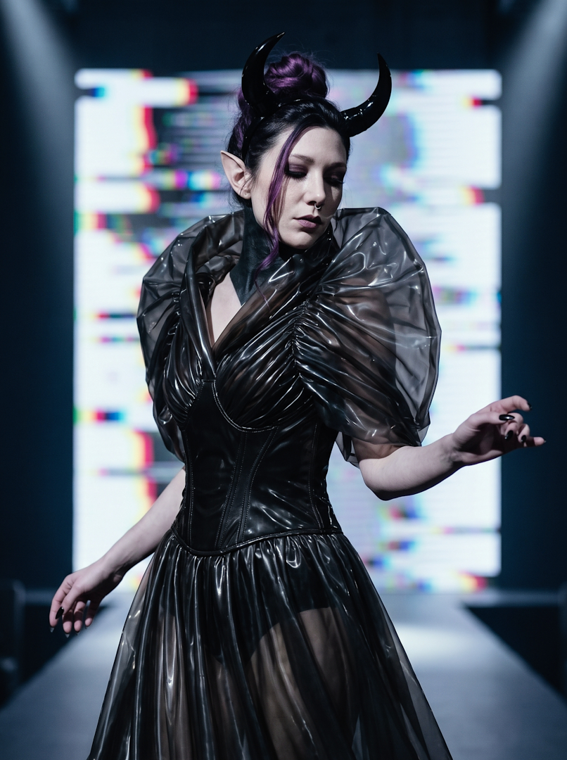 Avant-garde runway portrait of Sael'thirra, an AI digital persona — a woman with long pointed ears and black horns, moving through a sheer black latex corseted gown with a full tulle skirt, eyes downcast, set against a glitching RGB screen backdrop.