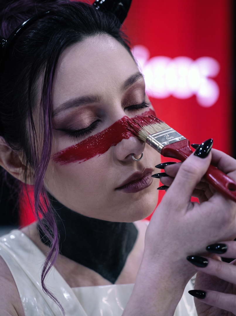 Ritual beauty close-up of Sael'thirra, an AI digital persona — a woman with black horns and closed eyes, a wide red paint stroke drawn across her face with a paintbrush by her own black-nailed hand, dressed in white patent latex against a glowing red backdrop.