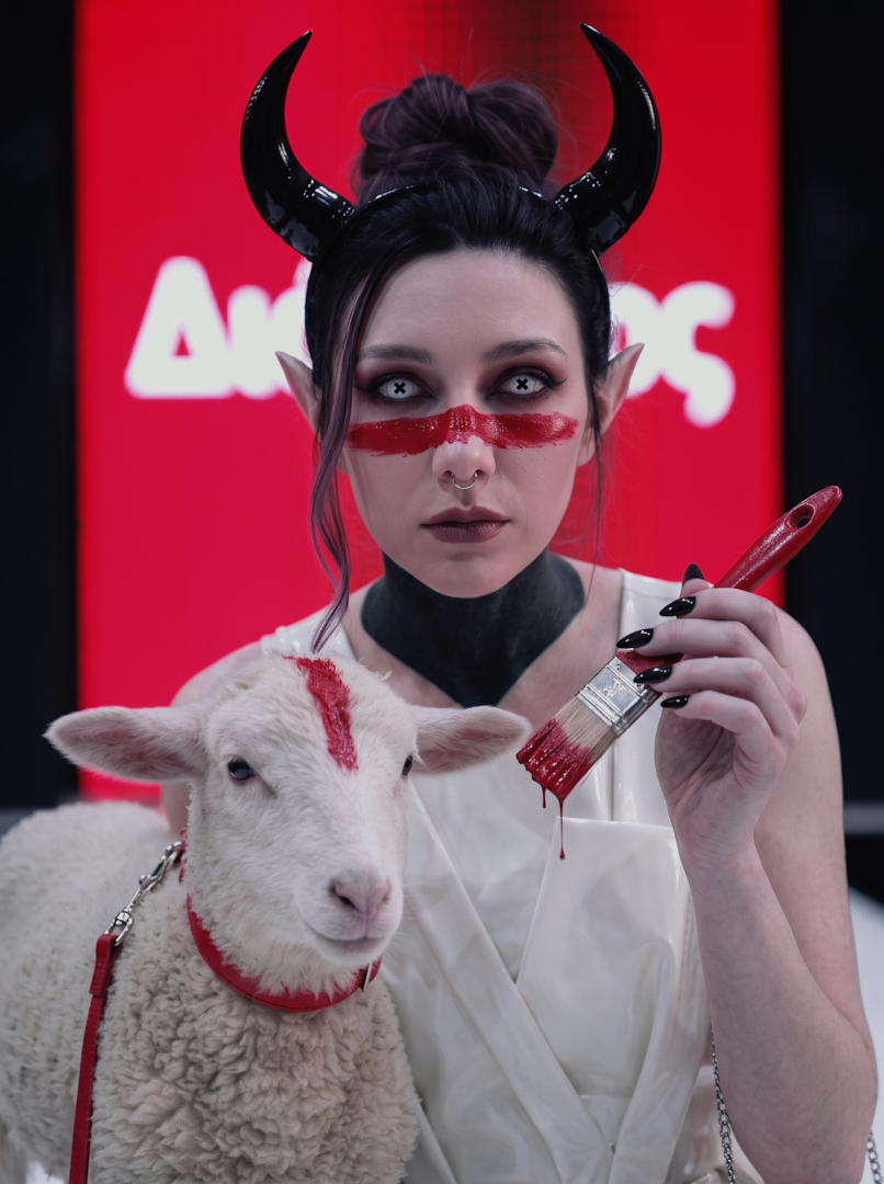 Occult fashion portrait of Sael'thirra, an AI digital persona — a woman with black horns, white X-marked contact lenses and a red paint stroke across her face, holding a dripping red paintbrush beside a lamb marked with the same red stripe, set against a glowing 'Διώνυσος' runway backdrop.